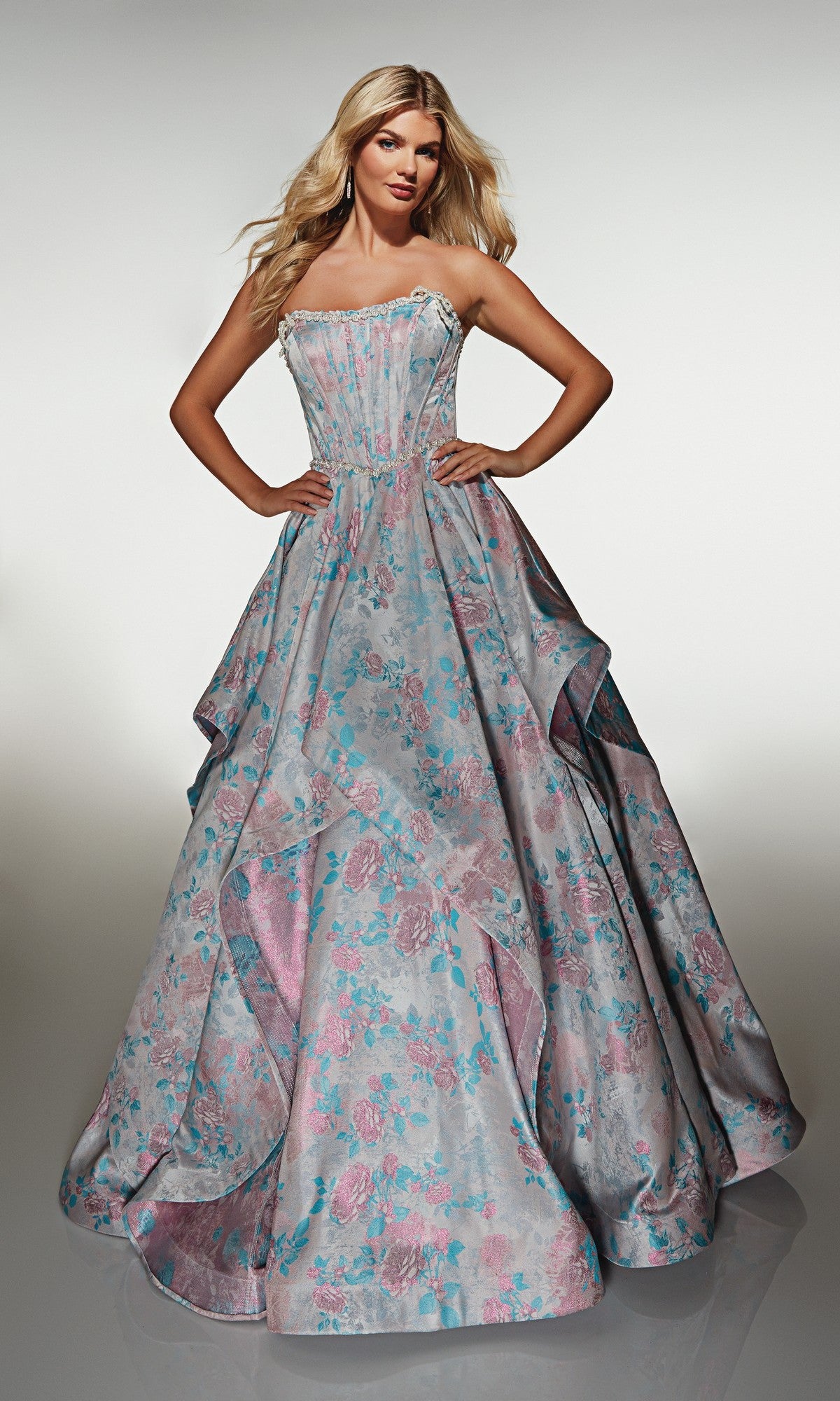 A woman stands with hands on hips against a plain background, wearing the Alyce 61997 Long Prom Dress—a strapless ball gown featuring a pastel blue and pink floral pattern and a corset bodice.